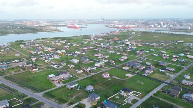 Homes along Seashell Dr sit south of Bluewater Highway, Freeport Harbor Channel visible beyond marsh corridor. Elevated roofs, sunlit grass contrast with cranes tank shadows in Freeport, TX