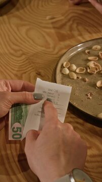 Vertical of hands of anonymous female customer checking receipt and counting US dollar banknotes on wooden table in bar while splitting bill with friends. Financial planning, budgeting, payment