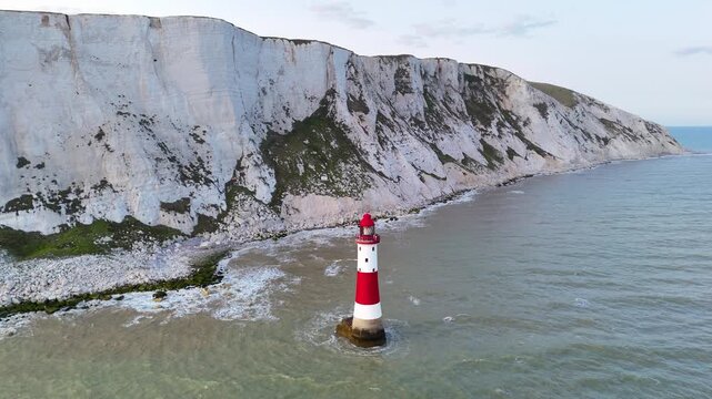 Drone footage approaching a red and white lighthouse surrounded by the sea with dramatic white chalk cliffs in the background on the southern coast of England.