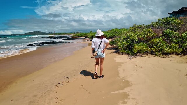 Woman walking along the coastline of Isabela Island, Galapagos. Back view of female traveler in white shirt and hat near volcanic rocks and green shrubs.