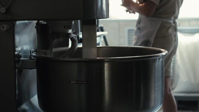 Female baker works near spiral dough mixer. Baker in professional kitchen, wearing apron, stands next to powerful spiral dough mixer. Mixing large batch of ingredients for bread making process. Focus 