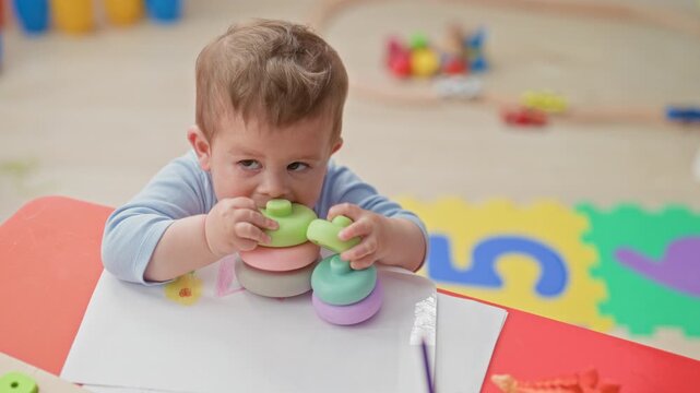 Boy toddler crying and gripping pastel stacking rings with both hands on a red play table beside foam number mat in building; upset.