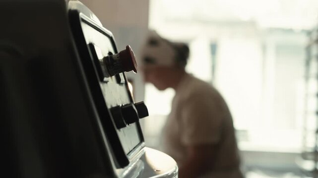 Female baker is working in background. Close up view electromechanical control panel with red emergency stop button for large spiral dough mixer. Dimly lit industrial kitchen background