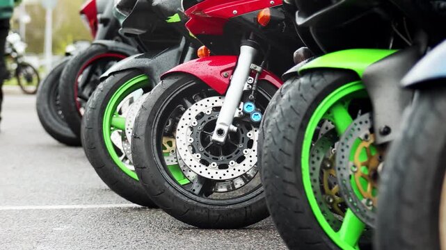 Close up view of several parked motorcycles lined up, featuring vibrant green and red fairings, highlighting suspension, tire treads, and disc brakes on a wet ground surface outdoors.