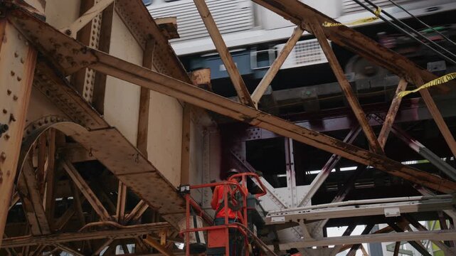Construction worker in boom lift grinding rusty steel girder of elevated Chicago L train track while public transit passes on bridge structure in city