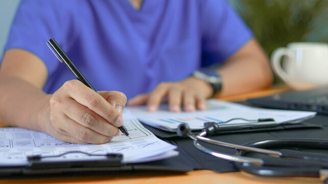 Healthcare professional reviewing medical documents and writing notes at a desk with stethoscope and laptop. Clinical documentation, patient records, healthcare administration, medical reporting