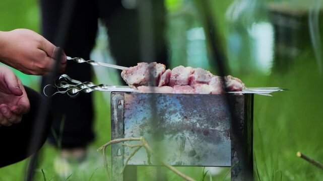 Close up view raw marinated meat pieces metal skewers over rustic metal brazier. Hot coals generate smoke heat for outdoor cooking. Person hand adjusts skewers, ensuring even cooking during summer gat