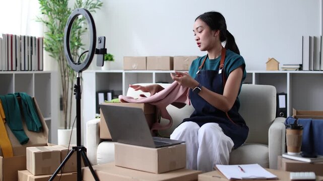 Asian businesswoman and small business owner live streaming on social media, using a smartphone and ring light to sell clothes online while working from her home office