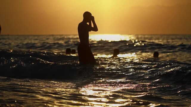 Man stands in ocean water, silhouetted against vibrant sunset sky, adjusting eyewear; several other people enjoying swim, golden reflections of sun on water create serene atmosphere. Vacation holiday 