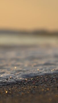 Detail shot of small waves with white foam reaching dark pebbles and sand on ocean shore. Warm golden light of sunset illuminates water and reflects off wet surface. Vacation holiday leave. Slow motio