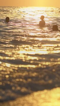 Silhouettes partially visible above ocean surface during golden hour. Sunlight reflects intensely off water creating dazzling bokeh. Gentle waves lap around figures, serene and picturesque natural sce