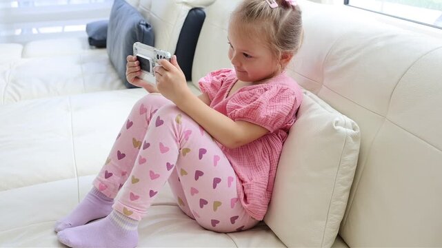 Young child concentrating on a smartphone screen while sitting indoors. Bright home interior, digital technology, entertainment and modern family lifestyle concept.
