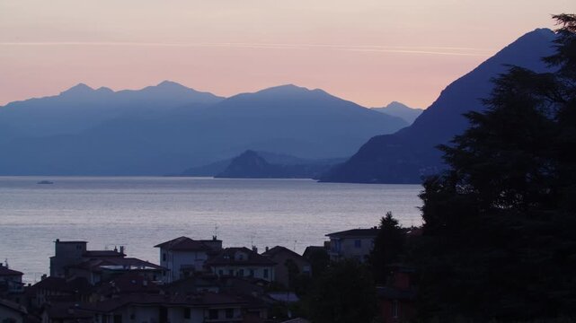 Wide panorama of Lake Maggiore at dawn from Stresa, Italy. Lakeside village silhouettes, alpine peaks, and quiet waters under a soft pink and purple morning sky.