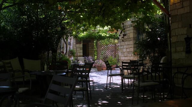 Traditional empty Greek tavern tables set outside under green trees during a bright summer day. Authentic rustic restaurant patio in Greece