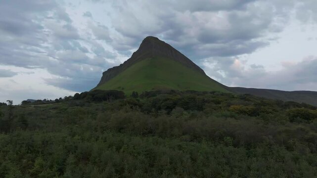 Cinematic aerial shot approaching Benbulbin mountain in Sligo, Ireland, with lush green forests in the foreground under a dramatic cloudy sky. Great for travel and nature documentaries.