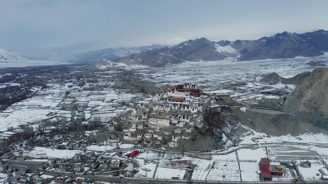 Thiksey Monastery sits atop a rocky hill surrounded by snow-covered agricultural fields in the Indus Valley. Himalayan mountain ranges frame the winter landscape under overcast skies.