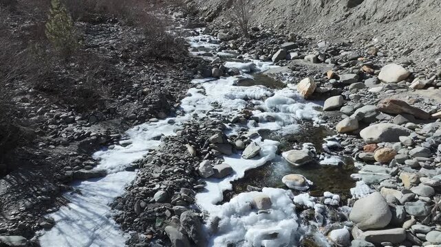 Mountain stream flows through ice and rocks in winter near Manali, Himachal Pradesh. Aerial view reveals the partially frozen waterway surrounded by barren slopes on Muling road near Sissu.