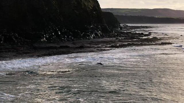 Slow motion aerial of ocean waves hitting the rocky shoreline at the base of dark cliffs in County Clare, Ireland. Authentic coastal landscape.