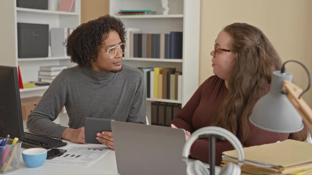 Man holding tablet shakes hand with woman across a laptop and paperwork at a desk in an office building; teamwork trust.