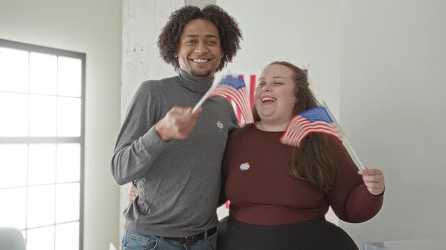 Man and woman smiling and waving american flags while wearing i voted stickers inside a civic building with window light; civic pride celebration.