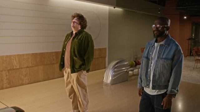 Diverse young friends, Caucasian man and African American man, throwing bowling balls and joking at missing pins in bowling alley. Ideal for lifestyle commercials, friendship, and leisure promos