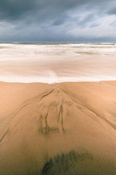 View of a sandy beach with water patterns and long exposure waves under a moody overcast sky in Debki, Pomeranian Voivodeship, Poland.