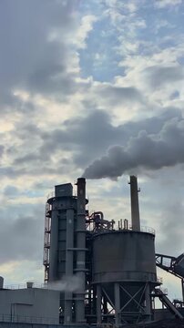Cement plant emitting smoke in low angle beside massive silos and conveyors for industrial production, concrete manufacturing, air pollution, energy use and heavy industry infrastructure workflow.