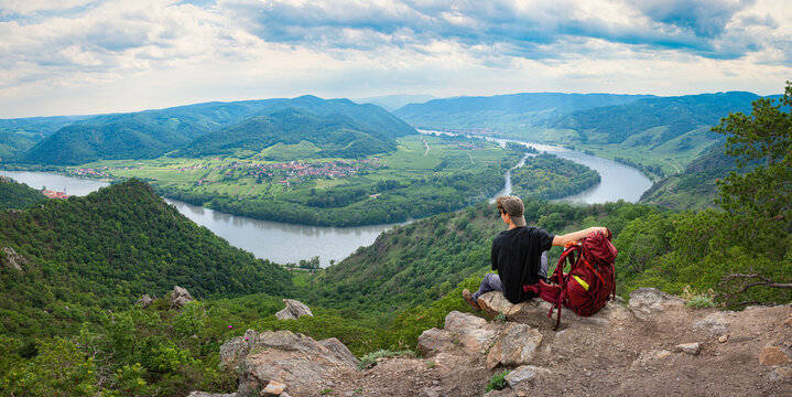 Hiker with backpack sitting on a rocky cliff looking at the panoramic view of the Danube river loop in Wachau valley, Austria