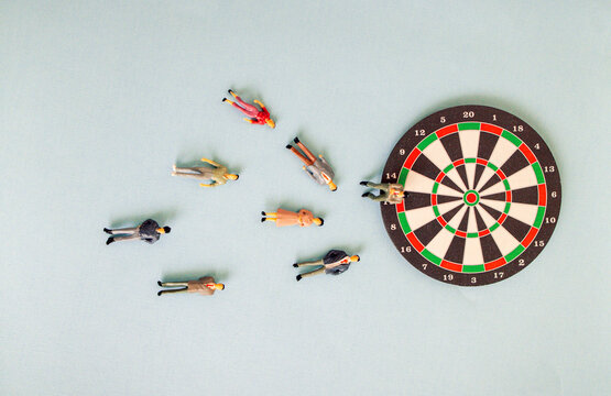 Miniature figurines scattered around a dartboard on a light blue background, depicting a playful scene of a dart game with various colorful figures