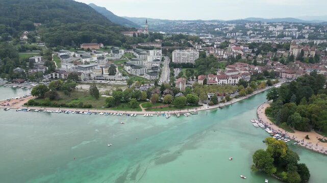 4K Aerial Drone View of Lake Annecy Waterfront Peninsula, Marina with Boats, and City of Annecy Surrounded by Green Hills and Forest Under Cloudy Sky, Haute‑Savoie, French Alps, France