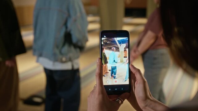 Hands of young woman holding smartphone and recording diverse friends throwing ball and dancing at bowling alley. Footage is ideal for social media marketing and mobile app advertisements