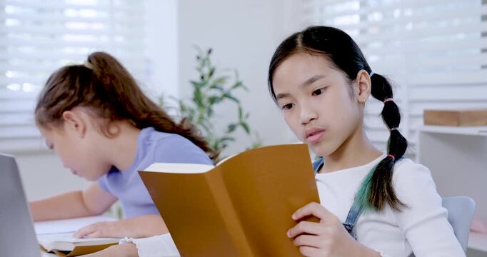 Asian girl preteen reading notebook carefully while caucasian girl preteen student concentrating on school assignment writing during classroom teamwork project digital education study collaboration