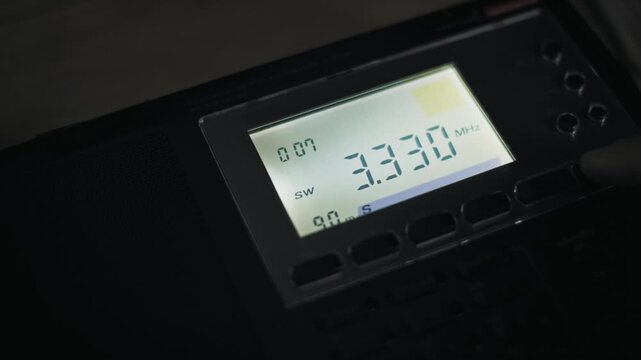 Dark scene of a finger pressing a button to tune a digital shortwave radio. The LCD screen backlight illuminates the changing MHz frequency and signal meter during the search.