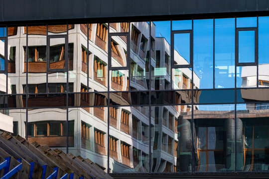 Layered urban architecture shows a glass facade reflecting office railing forms, with stair structure, window pattern, building depth and blue reflection across the frame