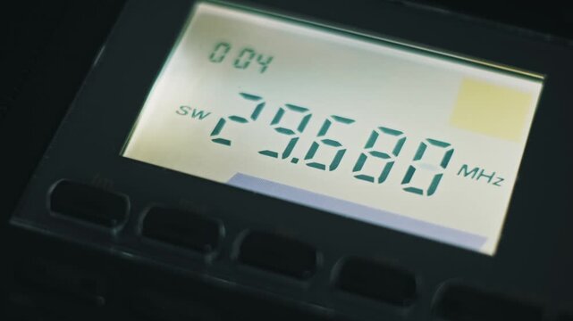 Macro close-up of a digital radio tuner scanning down through shortwave frequencies. The backlit LCD screen displays rapidly changing numbers with MHz units and a SW band indicator.