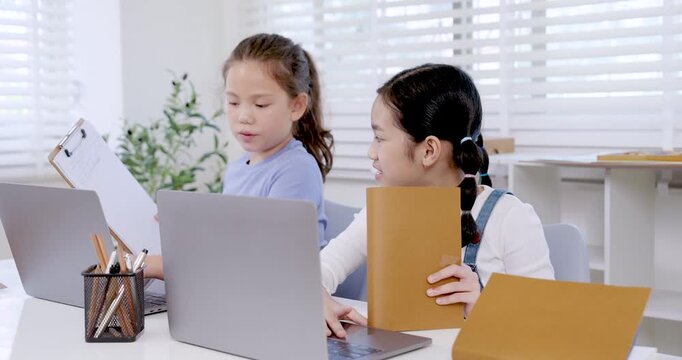 Asian preteen girl reading folder while caucasian girl watching beside laptop in classroom representing teamwork digital education collaboration learning project productivity study communication