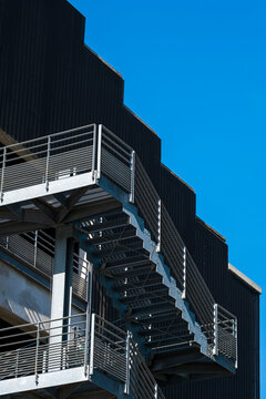 Metal stairs cut across black facade and blue sky in urban architecture, where railing, shadow, structure and exterior access form a sharp graphic angle