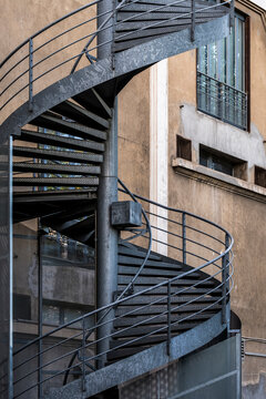 Spiral stairs climb beside metal railing and concrete wall in urban architecture, where shadow, exterior structure and industrial detail shape the frame