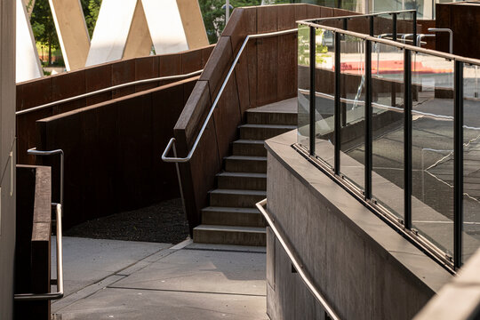 Urban architecture frames concrete stairs between metal railing, glass, walkway and structure, as sunlight and shadow lead through an exterior access route