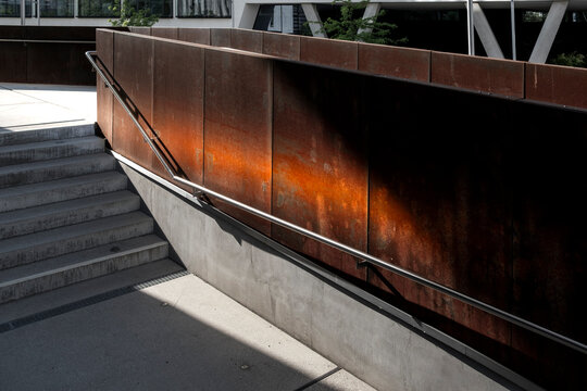 Concrete stairs pass a rusted metal railing in urban architecture, where sunlight, shadow, walkway, structure and exterior wall define a quiet access route