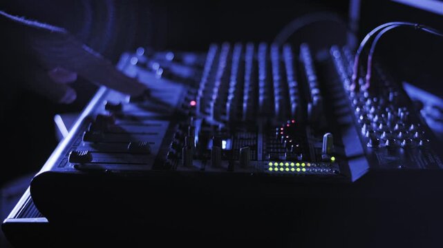 Medium shot of a person's hand adjusting various knobs and faders on an audio mixing console. Glowing LED level meters stand out in the dark, blue-lit music production space.