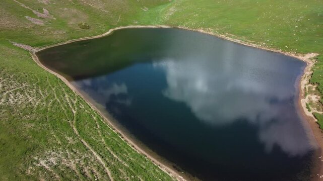 Drone birds eye view shot of a dark mountain lake resembling a heart shape, surrounded by green shores and a melting patch of snow in Sairam-Ugam National Park, Kazakhstan. 