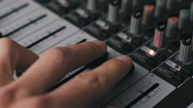 Angled close-up producer's hand adjusting two faders simultaneously on a sound board. The shot features cool lighting, shallow depth of field, and multiple illuminated buttons in a studio.