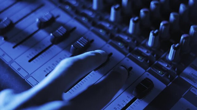 Detailed view of fingers sliding multiple faders on a professional soundboard. Decibel markers and control knobs are visible under shifting blue and purple stage lighting.