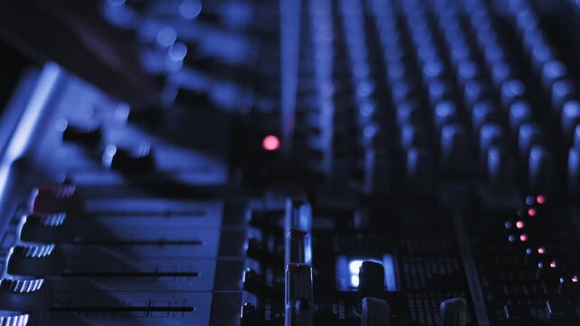 Side angle of a sound engineer's hand moving faders on a mixing console to adjust audio levels. The scene features blinking lights and a shallow depth of field under cool blue lighting.