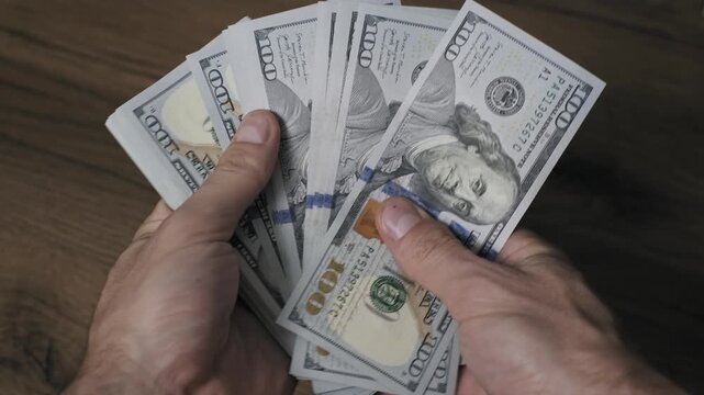 Close-up of male hands fanning out a thick stack of one hundred dollar bills over a dark wooden table. This display of hard cash represents wealth, profit, and financial success.