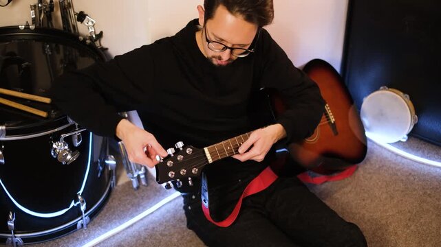 Young handsome guitarist in glasses tuning guitar sitting in his studio

