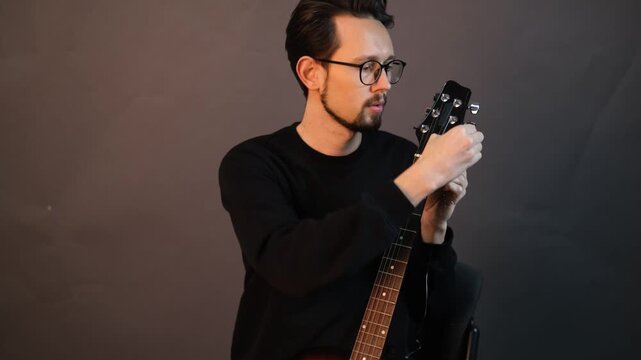 Young handsome talented guitarist in glasses replacing strings on guitar tuning it

