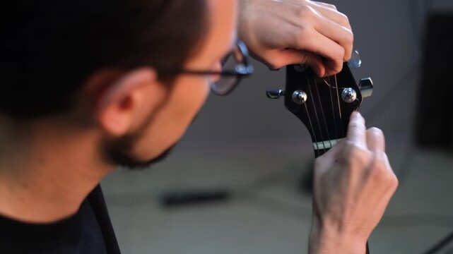 Young handsome talented guitarist in glasses replacing strings on guitar tuning it


