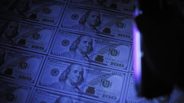 Angled perspective of a UV light beam scanning rows of hundred dollar bills on a dark surface. The silhouette of the lamp moves to reveal hidden watermarks for a counterfeit check.